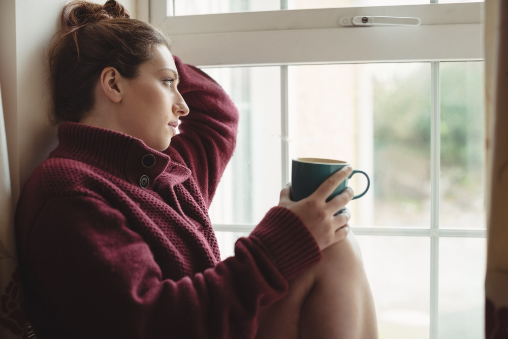 Mujer sentada mirando por la ventana con una taza de café, simbolizando descanso mental, reducción del cansancio y recuperación de la motivación