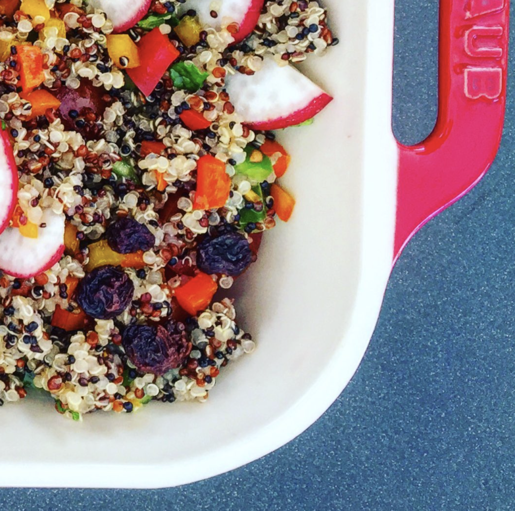 Detalle de ensalada de quinoa tricolor con uvas pasas, rábano, pimientos y hierbas frescas, servida en bol blanco con vinagreta ligera.